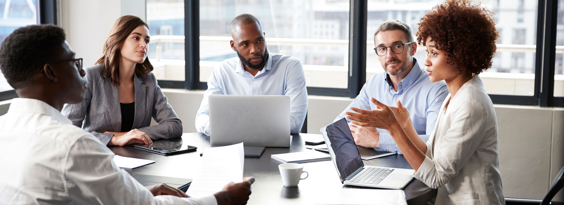 In a spacious and bright boardroom, a diverse group of professionals listen intently as their coworker speaks