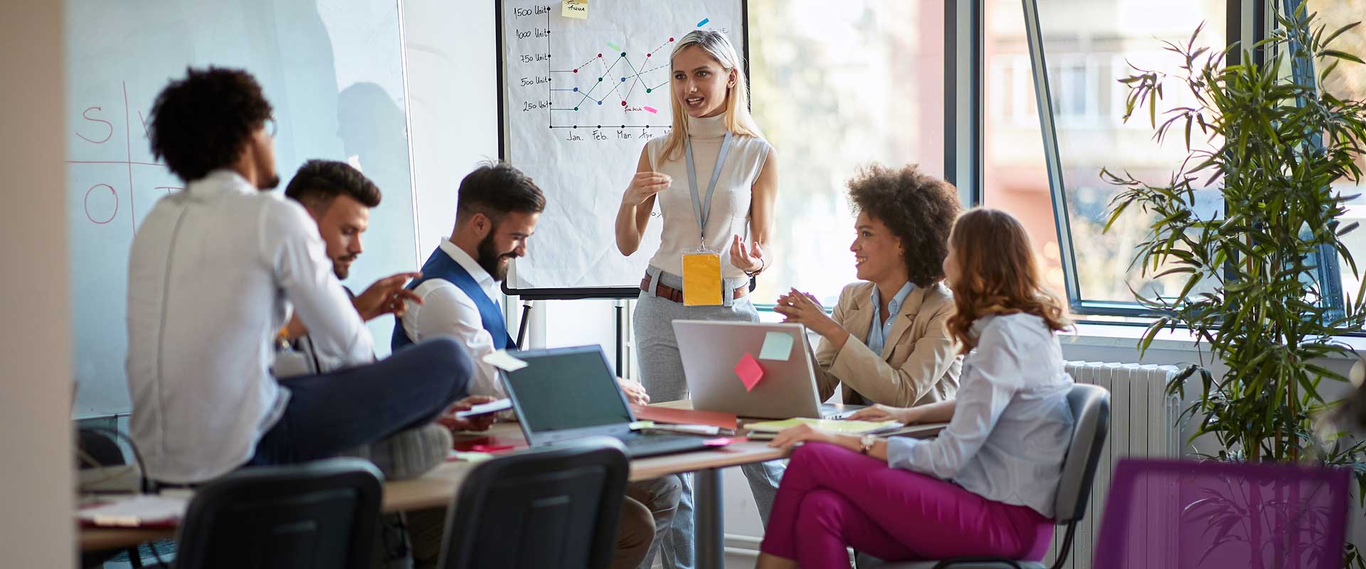 In a bright boardroom with large windows, a woman gives a presentation to her colleagues